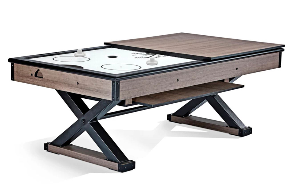 A modern air hockey table with a wooden finish, black metal legs in an X shape, and a partially covered top that doubles as a tabletop. Two white strikers and a puck are visible on the playing surface.